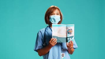 Experienced nurse presenting medical leaflet containing list of clinic professional services. Hospital representative advertising healthcare brochure, isolated over blue studio background photo