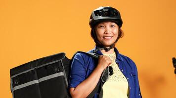 Portrait of happy asian courier using bike and thermic bag to transport perishables to customer. Delivery part time employee isolated over orange studio background wearing safety gear, close up photo