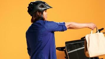 Close up of delivery bike rider greeting customer, offering takeout meal bag, isolated over studio background. Asian courier woman reaching food order location, waiting for client to answer door photo