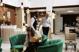 Friendly african american bellhop assists senior man in hotel lobby with luggage to the reserved room. Elderly female visitor reads while waiting for accomodation in cosy lounge area with armchairs. photo