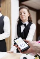 Female receptionist at front desk providing assistance to tourist using mobile phone for nfc payment. Image showcasing hotel concierge holding pos terminal for traveler to pay with smartphone. photo