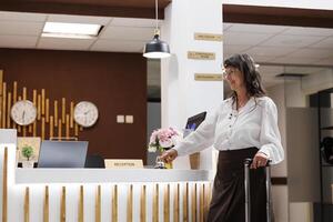 Senior woman reaches registration counter and rings the metallic service bell asking for services from hotel receptionists. Caucasian elderly female tourist awaiting assistance from resort personnel. photo