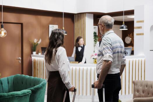 Retired senior couple holding trolley bags entering hotel reception, ready for fun honeymoon vacation. Elderly male and female travelers approaching front desk to speak with receptionists. photo