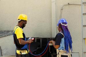 African american coworkers hired after complaints about noisy air conditioner and significant increase in energy bills. Engineers using professional manifold gauges to detect optimum fixing method photo