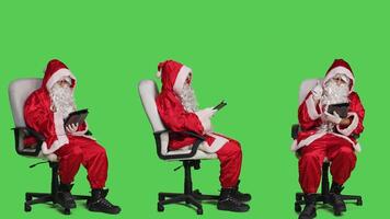 Modern person using digital tablet during christmas eve holiday celebration, checking online webpage while he sits on chair in studio. Santa in suit on full body greenscreen backdrop. photo