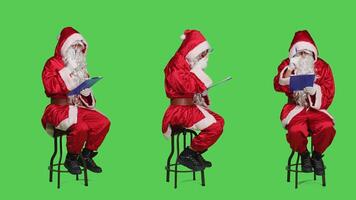 Saint nick writing notes on clipboard while he sits on chair dressed in red costume and white beard, full body greenscreen. Father christmas character holding papers in studio. photo