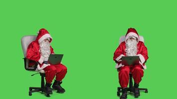 Modern saint nick works on laptop in studio, sitting on chair against full body greenscreen background. Santa claus character using wireless portable pc, browsing online site in festive costume. photo