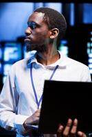 African american manager in server hub using laptop to find firewall misconfigurations affecting rackmounts performance and connectivity, leading to sluggish data transfer rates photo