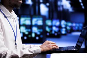 African american supervisor in server room using laptop to set up configuration management tools that enable automatic failover and load balancing, preventing high tech equipment data loss photo