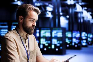 System administrator solving performance related issues in server cabinets. Expert utilizing tablet to identify operational problems causing high tech workplace infrastructure slowdown photo