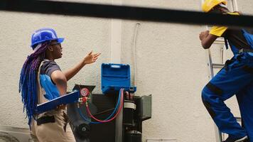 Precise worker measuring refrigerant levels in air conditioner with professional manometers while coworker steps down from folding ladder after finishing checking rooftop HVAC system photo