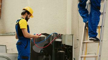 Expert mechanic measuring refrigerant levels in air conditioner with manifold gauges while african american coworker steps down from folding ladder after finishing checking rooftop HVAC system photo