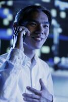 Cheerful man strolling around town during nighttime, enjoying discussion over the phone with girlfriend, close up. Asian citizen making telephone calls while commuting on dimly illuminated streets photo