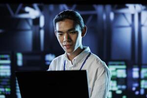 Asian man between server hub rows providing processing resources for businesses worldwide. System administrator fixing data center mainframes tasked with managing massive databases photo