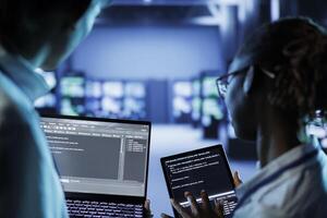 IT developers building firewall systems in server room housing advanced infrastructure. Licensed specialists running code on tablet and laptop, to upgrade data center rigs handling demanding workloads photo