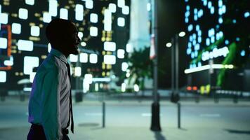 africano americano persona en noche caminar céntrico debajo luces de la calle, admirativo moderno oficina edificios desde iluminado acera. confidente hombre disfrutando Noche paseo alrededor ciudad. foto