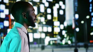 Businessman admiring skyscrapers at night, walking around city center with streetlights. African american person feeling happy on urban downtown promenade, relaxing on sidewalk. Handheld shot. photo