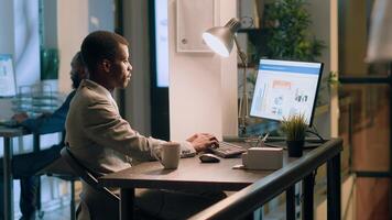 Portrait of african american computer operator sitting at desk during nightshift, looking through online financial business documents while coworker in background goes to bathroom photo
