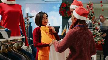 Asian retail assistant and coworker helping indecisive customer choose between two colors of blazers in Christmas decorated clothing shop. Employees assisting client during winter holiday season photo