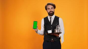 Bearded waiter presents greenscreen on phone display, holding smartphone to show isolated mockup screen. Elegant skilled restaurant valet using empty copyspace template in studio. photo