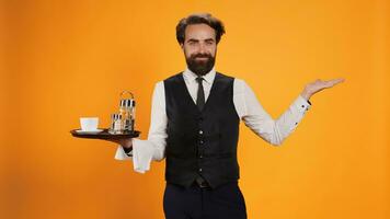 Smiling waiter posing with food tray in hand, carrying utensils and coffee cup to serve customers against yellow background. Confident employee working at five star restaurant, taking meal orders. photo