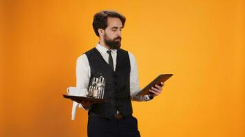 Personnel serving coffee order and holding tablet in studio. Posing in front of yellow background, experienced stylish butler preparing to give bill to customers using electronic device. photo