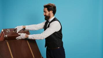 Classy hotel employee with baggage posing against blue background to advertise tourism or travel field. Bellboy working in hospitality with formal clothing attire, carrying trolley bags. photo