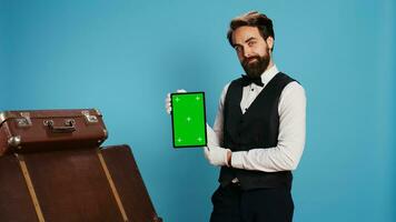 Doorman poses with greenscreen tablet on camera, standing next to blue background in studio. Hotel worker showcasing isolated mockup copyspace on device, hospitality concept. photo