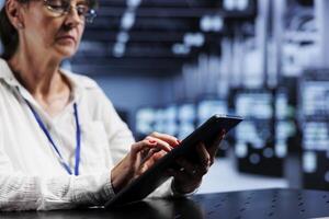 Aged technical support consultant using tablet to assess server security glitches arising due to unsecured network connections. Certified expert ensuring data center is impenetrable by virus attacks photo