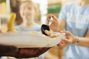 Photo focus on caucasian woman serving baked beans to hungry underprivileged african american person at a non-profit food drive. Close-up shot of free food distribution by humanitarian aid team.