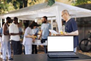 Picture shows digital laptop with a blank white screen that is sitting on a table while charity workers help the less fortunate. Minicomputer in the foreground with isolated copyspace mockup template. photo