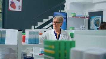 Aged knowledgeable pharmacologist in dispensary holding shopping basket, arranging merchandise on shelves. Older pharmacist organizing medicinal products in drugstore with customers in background photo