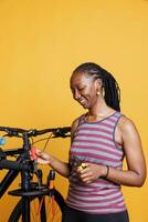 enfocado africano americano hembra ciclista expertamente refacción y mantiene un moderno bicicleta utilizando un rango de herramientas. sonriente negro mujer arreglando profesional equipo para fijación dañado bicicleta. foto