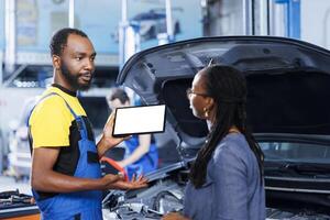 Qualified serviceman in car service uses mockup tablet to order new exhaust pipes for broken vehicle. BIPOC garage specialist shows client needed components replacement on isolated screen device photo