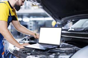 Mockup laptop on car with hood open while mechanic in blurry background replaces steering mechanism. Isolated screen device next to precise garage worker mending client vehicle photo