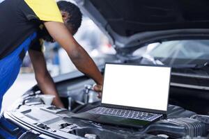 Mockup laptop on car with hood open while BIPOC repairman in blurry background replaces brakes. Isolated screen device next to experienced garage worker fixing customer automobile photo