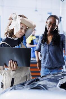 Technician in car service uses laptop to calculate invoice for client after repairing compressor belt. Tired garage employee calculating final costs after servicing customer vehicle photo