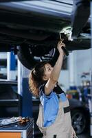Seasoned mechanic working on suspended car in garage, checking engine during routine maintenance. Auto repair shop professional underneath vehicle, inspecting components using work light photo