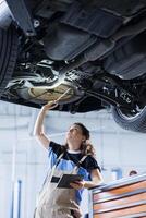 Woman working on suspended car in garage, checking tires during routine maintenance. Experienced auto repair shop expert underneath vehicle, inspecting it using work light photo