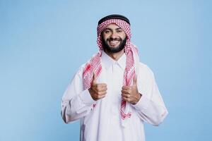 Muslim man in white thobe and checkered headdress posing with thumbs up showing approval studio portrait. Smiling arab person wearing traditional clothes looking at camera photo