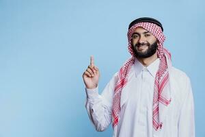 Young arab man in thobe and ghutra promoting product by pointing upwards with finger studio portrait. Muslim person advertising and showing up while looking at camera with cheerful expression photo