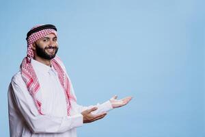 Cheerful muslim man in traditional clothes showing with hands at copy space for advertisement studio portrait. Smiling arab person showcasing to side with arms and looking at camera photo