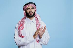 Confident muslim man in traditional attire applauding, displaying pride studio portrait. Arab peron dressed in white thobe clapping hands, showing support and looking at camera photo