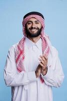 Excited muslim man in thobe and ghutra standing confidently, applauding with hands studio portrait. Smiling arab person clapping arms at performance and looking at camera photo