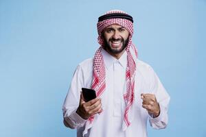 Happy arab reading good news on mobile phone and looking at camera with excited expression. Cheerful muslim man in thobe using smartphone and showing winner gesture with clenched fist portrait photo