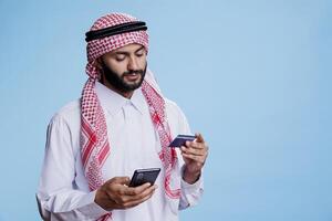 Muslim man wearing thobe using credit card and mobile phone while making electronic payment. Arab in traditional clothes holding smartphone while engaging in ecommerce shopping photo