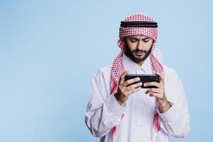 Arab man dressed in traditional attire enjoying leisure activity while playing video game on smartphone. Muslim person engaging in online gaming using mobile phone application photo