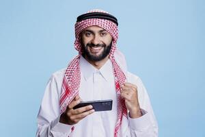 Happy muslim man playing online game on smartphone and showing winner concept studio portrait. Arab person winning videogame and looking at camera with clenched fist and excited expression photo