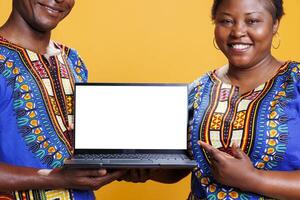 Cheerful black couple holding laptop together and pointing with finger at blank white screen. Smiling man and woman presenting portable computer with empty display with copy space photo