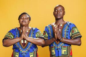 Black man and woman with closed eyes standing with folded palms, praying and asking for blessing. African american couple wearing ethnic clothes meditating and relaxing together photo